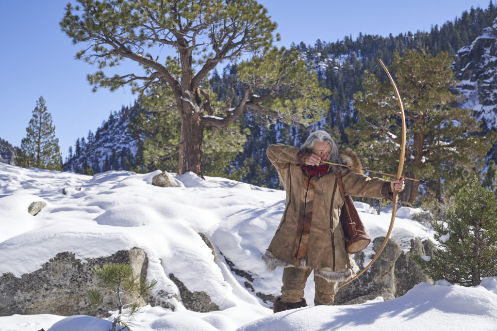 Woniya Thibeault (@buckskin_revolution) stands on a snowy mountain with her bow and arrow drawn.