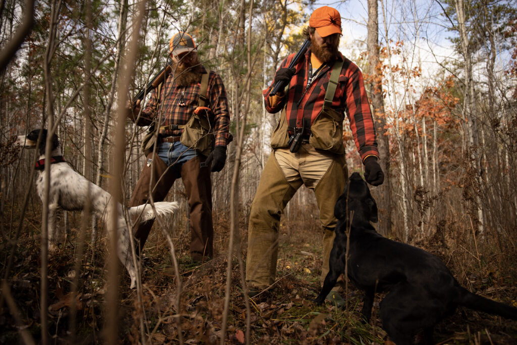 Two men stand beside two dogs in the woods during fall, both holding hunting rifles and dressed in hunting gear.