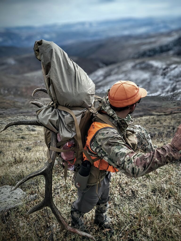 Casey Gorsett facing a mountain wearing a backpack in a orange Stormy Kromer.