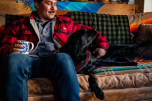 Man sits on a couch with a mug of coffee and hugs his black lab.